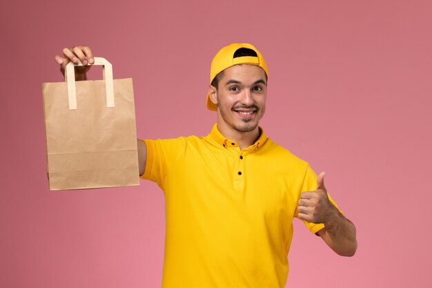 Front view male courier in yellow uniform holding delivery paper package smiling on light pink background.