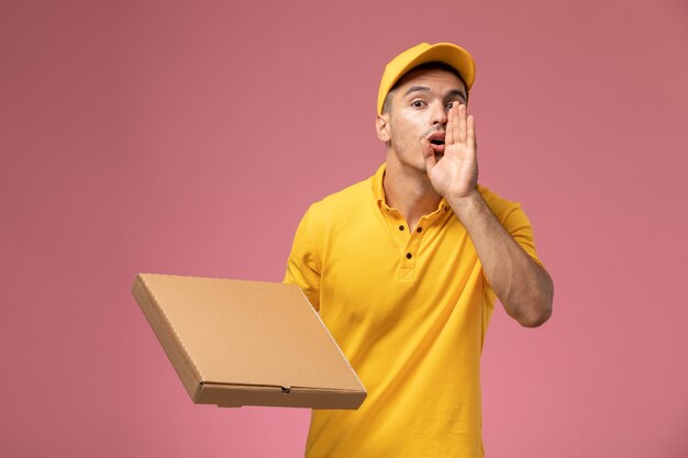 Front view male courier in yellow uniform holding food delivery box and calling out someone on the pink background 