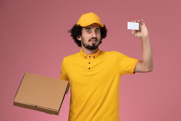 Курьер-мужчина, вид спереди в желтой форме, держит коробку для доставки еды и открытку на розовой стене