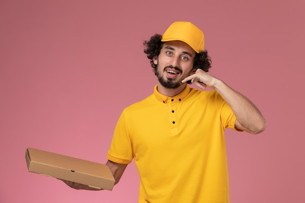 Front view male courier in yellow uniform holding food delivery box on light-pink wall