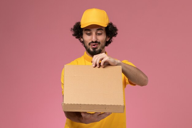 淡いピンクの壁に食品配達ボックスを保持している黄色の制服を着た正面図の男性宅配便