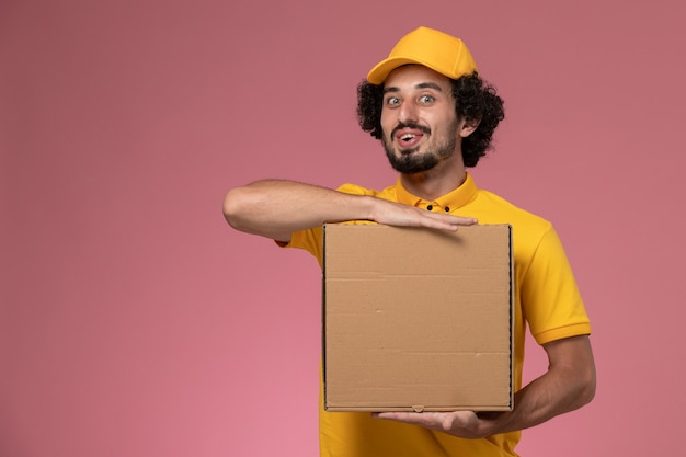 Front view male courier in yellow uniform holding food delivery box on the light-pink wall