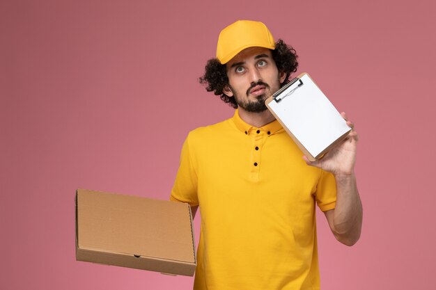 Курьер-мужчина, вид спереди в желтой форме, держит коробку для доставки еды и блокнот, глубоко задумавшись на розовой стене