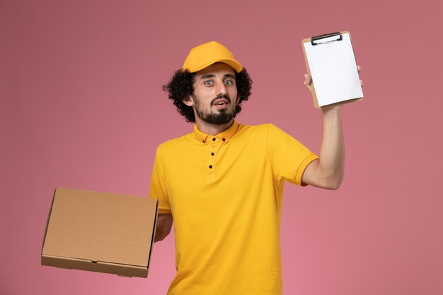 Front view male courier in yellow uniform holding food delivery box and notepad on light pink wall