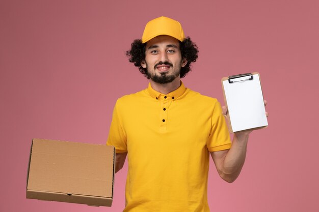 Front view male courier in yellow uniform holding food delivery box and notepad on pink wall