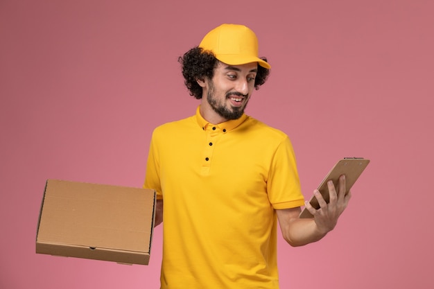 ピンクの壁に笑みを浮かべて食品配達ボックスとメモ帳を保持している黄色の制服を着た正面図男性宅配便