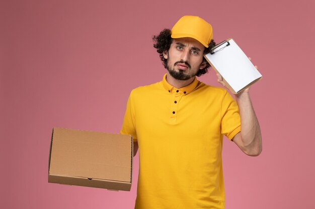 Front view male courier in yellow uniform holding food delivery box and notepad thinking on pink wall