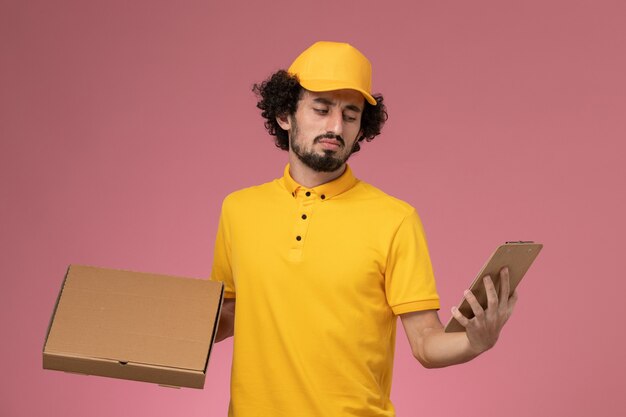 Front view male courier in yellow uniform holding food delivery box and notepad thinking on pink wall