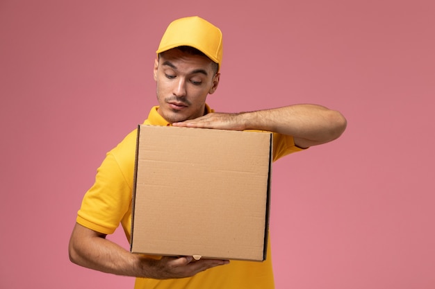 Курьер-мужчина в желтой форме, держащий коробку для доставки еды на розовом столе, вид спереди
