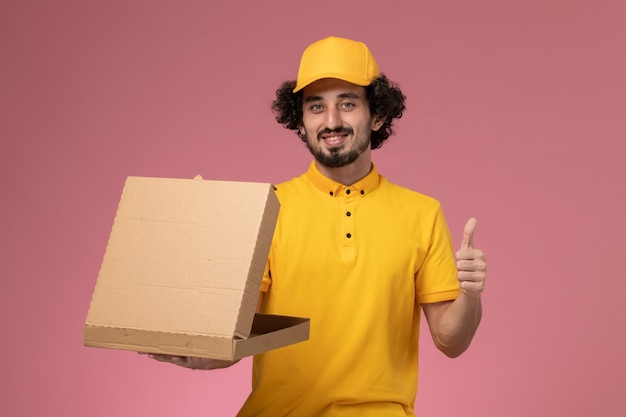 Front view male courier in yellow uniform holding food delivery box smiling on the light-pink wall