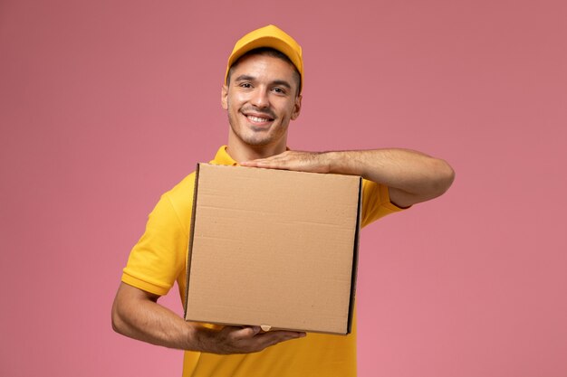 Front view male courier in yellow uniform holding food delivery box and smiling on the pink background 