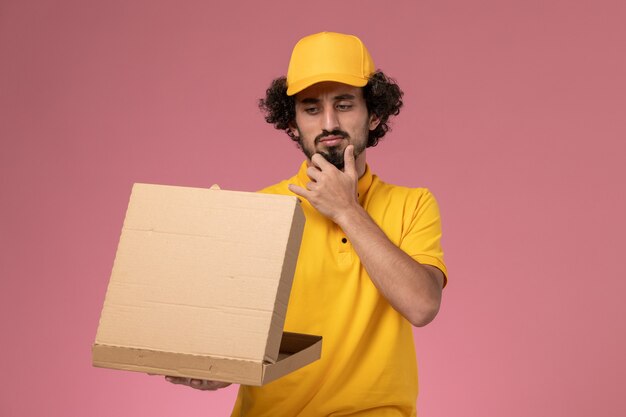 Front view male courier in yellow uniform holding food delivery box thinking on the light-pink wall