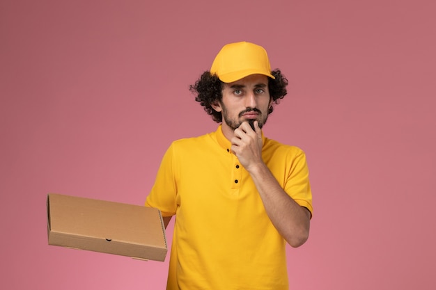 Front view male courier in yellow uniform holding food delivery box thinking on the light-pink wall