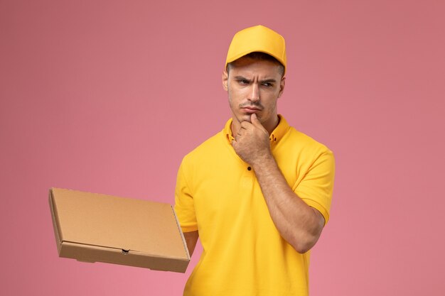 Курьер-мужчина в желтой форме, держащий коробку для доставки еды и думающий на розовом фоне, вид спереди