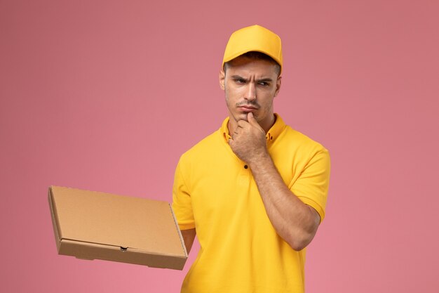 Front view male courier in yellow uniform holding food delivery box and thinking on the pink background 