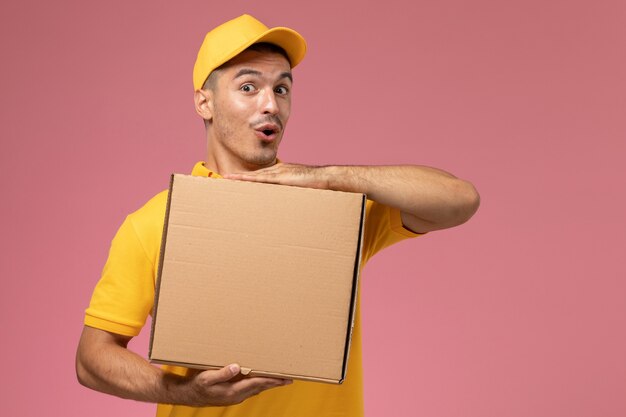 Front view male courier in yellow uniform holding food delivery box with funny expression on the pink desk 