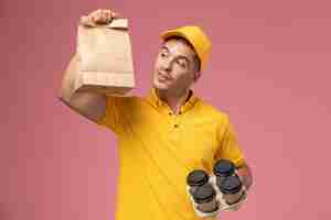 Free photo front view male courier in yellow uniform holding food package and delivery coffee cups on light-pink background