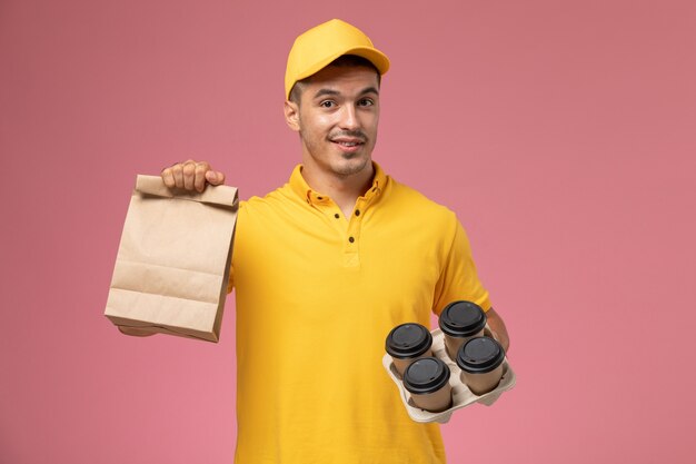 Front view male courier in yellow uniform holding food package and delivery coffee cups on the pink background