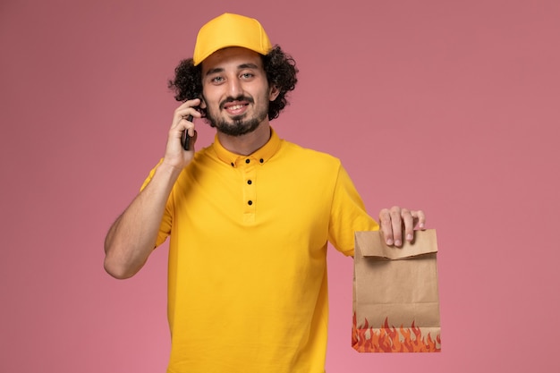 ピンクの壁に電話で話している食品パッケージを保持している黄色の制服を着た正面図男性宅配便