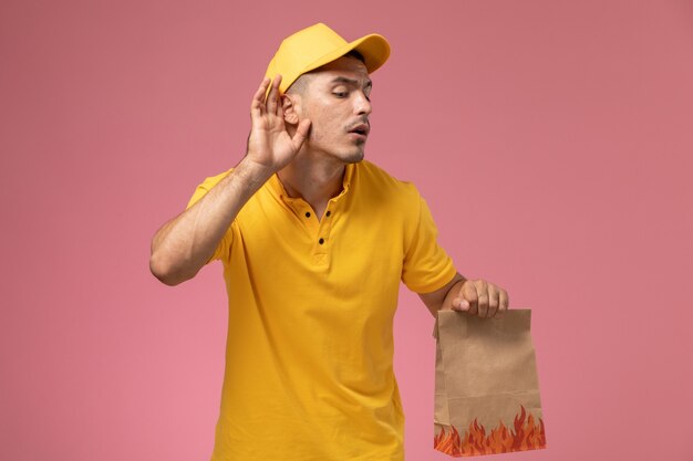 Front view male courier in yellow uniform holding food package trying to hear out on the pink background  