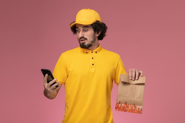 Front view male courier in yellow uniform holding food package and using phone on the pink wall