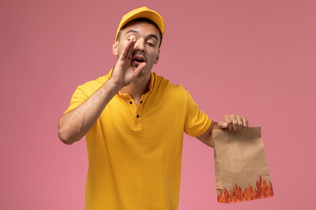 Front view male courier in yellow uniform holding food package whispering on the pink background  