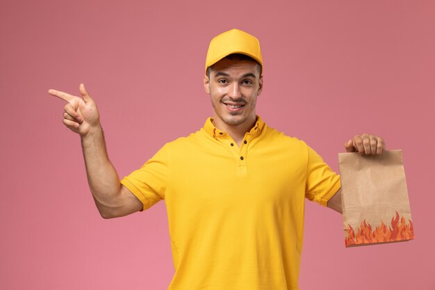 Front view male courier in yellow uniform holding food package with slight smile on pink background  