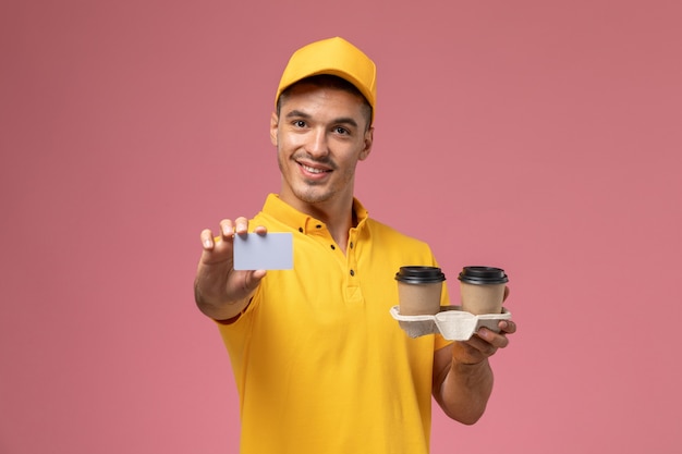 Front view male courier in yellow uniform holding grey card and delivery coffee cups on light-pink background 