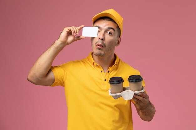 Front view male courier in yellow uniform holding grey card and delivery coffee cups on pink background