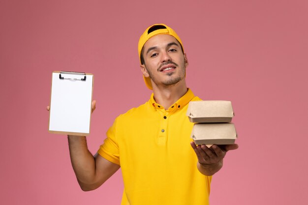 Front view male courier in yellow uniform holding little food packages on pink background.