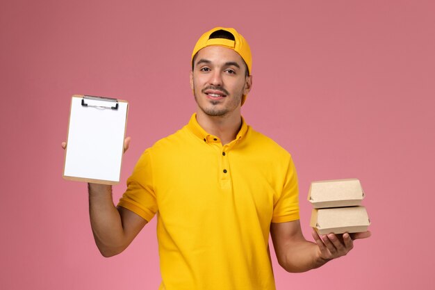 Front view male courier in yellow uniform holding little food packages on pink background.