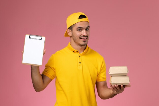 Front view male courier in yellow uniform holding little food packages on pink background.