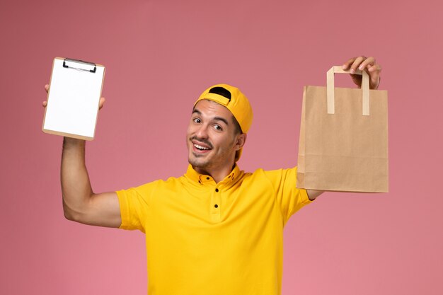 Front view male courier in yellow uniform holding little notepad and delivery food package on the light-pink background.