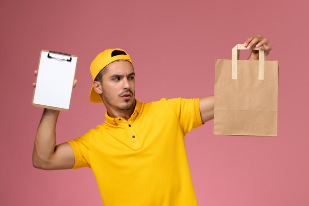 Front view male courier in yellow uniform holding little notepad and delivery food package on pink background.