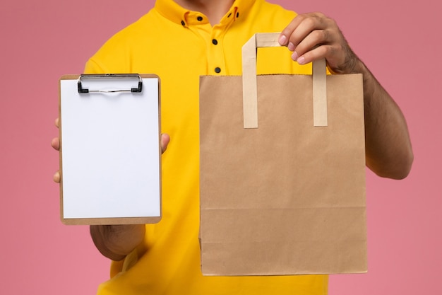 Foto gratuita corriere maschio di vista frontale in uniforme gialla che tiene piccolo blocco note e pacchetto di cibo di consegna sullo sfondo rosa.