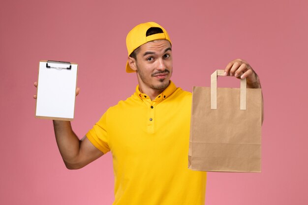 Курьер мужского пола вида спереди в желтой форме держа маленький блокнот и пакет еды доставки на розовом фоне.