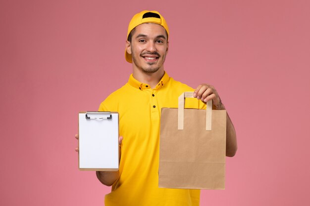 Front view male courier in yellow uniform holding little notepad and delivery food package on the pink background.