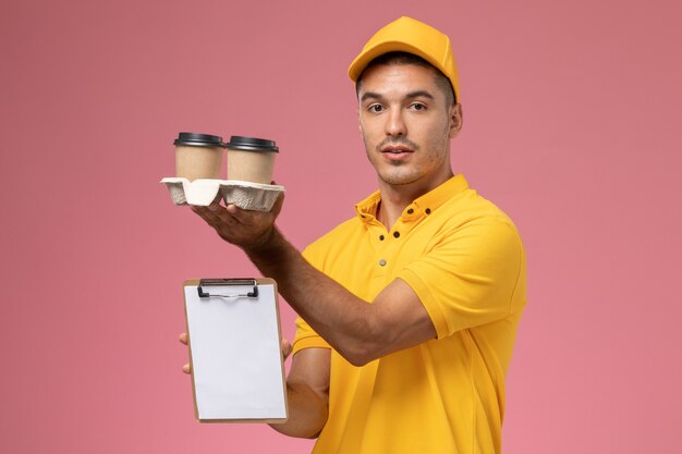 Front view male courier in yellow uniform holding notepad and delivery coffee cups on pink background