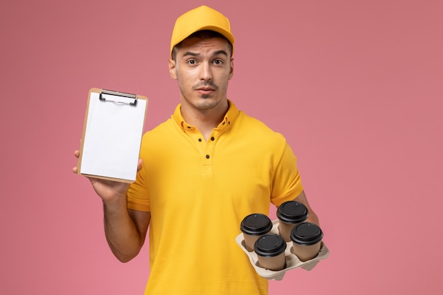 Front view male courier in yellow uniform holding notepad and delivery coffee cups on pink desk  