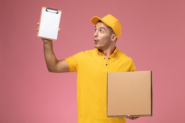 Front view male courier in yellow uniform holding notepad and food delivery box on the pink background   