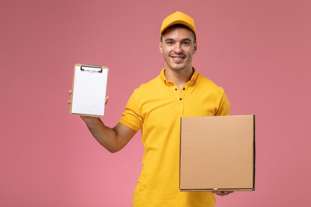 Front view male courier in yellow uniform holding notepad and food delivery box with smile on pink background   