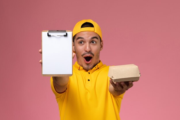 ピンクの机の上にメモ帳と小さな食品パッケージを保持している黄色の制服を着た正面図の男性宅配便。