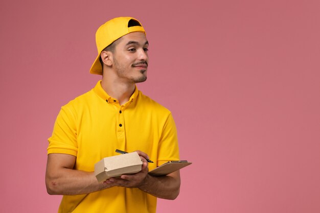 ピンクの背景にメモ帳と小さな食品パッケージを保持している黄色の制服を着た正面図の男性宅配便。