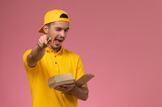 Front view male courier in yellow uniform holding notepad and little food package writing notes winking on pink background.