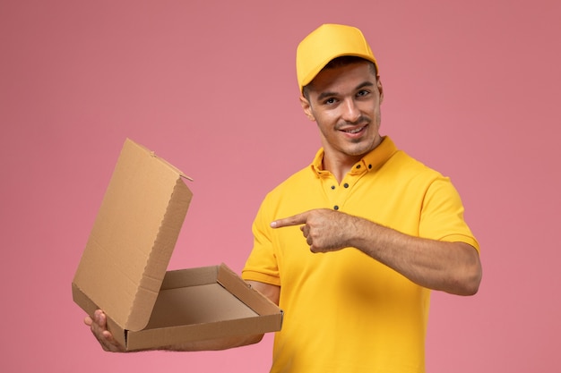 Front view male courier in yellow uniform holding and opening empty food delivery box on the pink desk 