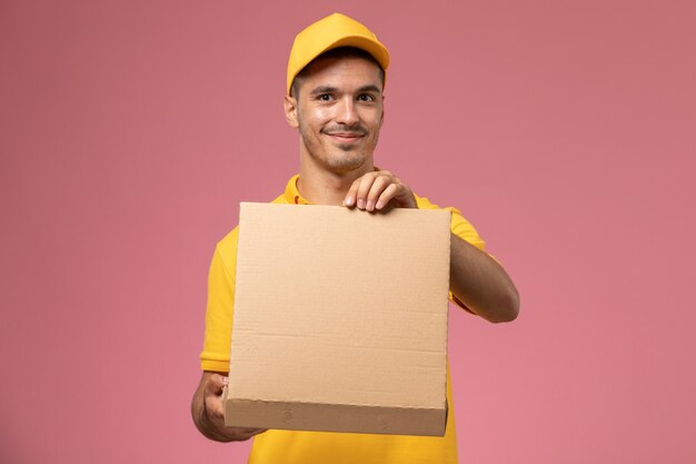 Front view male courier in yellow uniform holding and opening food delivery box on pink background 