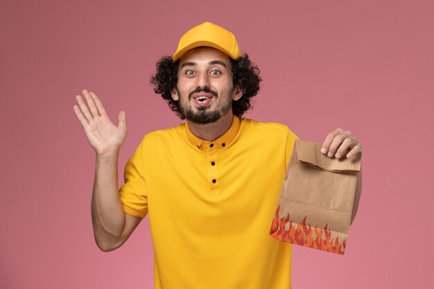 淡いピンクの壁に紙の食品パッケージを保持している黄色の制服の正面図男性宅配便