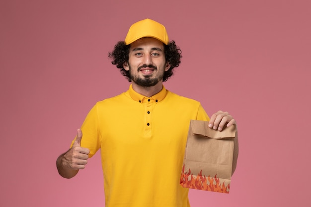 Front view male courier in yellow uniform holding paper food package on light pink wall