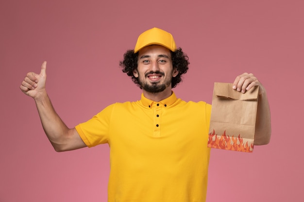 Front view male courier in yellow uniform holding paper food package on light pink wall