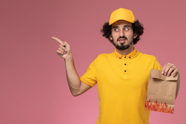 淡いピンクの壁に紙の食品パッケージを保持している黄色の制服の正面図男性宅配便